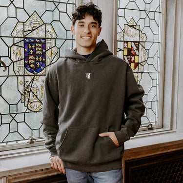 College-aged man wearing the Theta Chi embroidered black hoodie, standing in front of stained glass windows with fraternity-style crest designs.