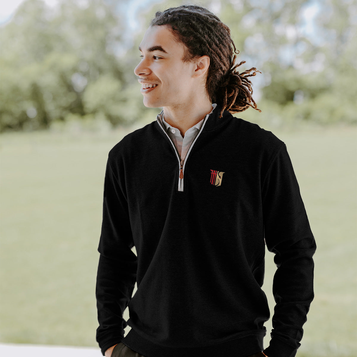 College-aged man smiles and looks off to the side while wearing a black quarter-zip pullover embroidered with a fraternity symbol. He stands outdoors in a bright, grassy setting.