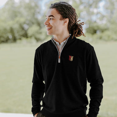 College-aged man smiles and looks off to the side while wearing a black quarter-zip pullover embroidered with a fraternity symbol. He stands outdoors in a bright, grassy setting.