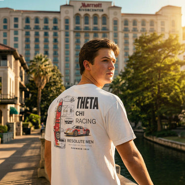 Rear view of a male model wearing the Theta Chi Racing t-shirt, looking left on an urban riverwalk with a hotel in the background.
