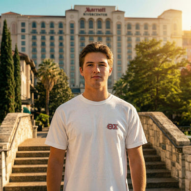 Front view of the male model wearing the Theta Chi t-shirt, showing the chest logo, standing on stone stairs by a canal with a hotel in the distance.