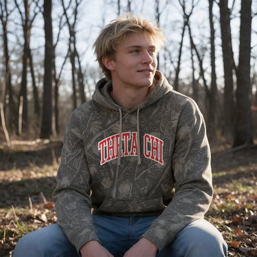 College-aged male wearing the fraternity Deadwood Camo Hoodie with red and white Theta Chi lettering, seated outdoors in a wooded setting.