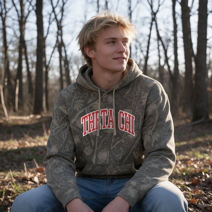 College-aged male wearing the fraternity Deadwood Camo Hoodie with red and white Theta Chi lettering, seated outdoors in a wooded setting.