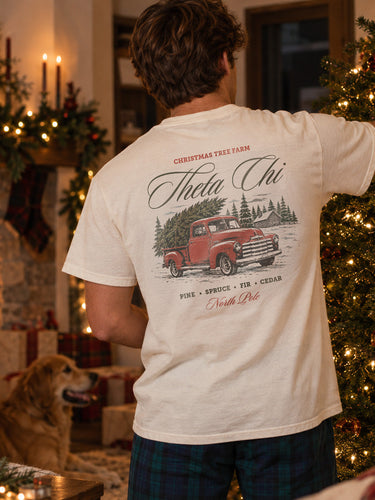 College-aged man wearing a limited edition Tree Farm Holiday Tee in a cozy Christmas setting with a decorated tree and fireplace.
